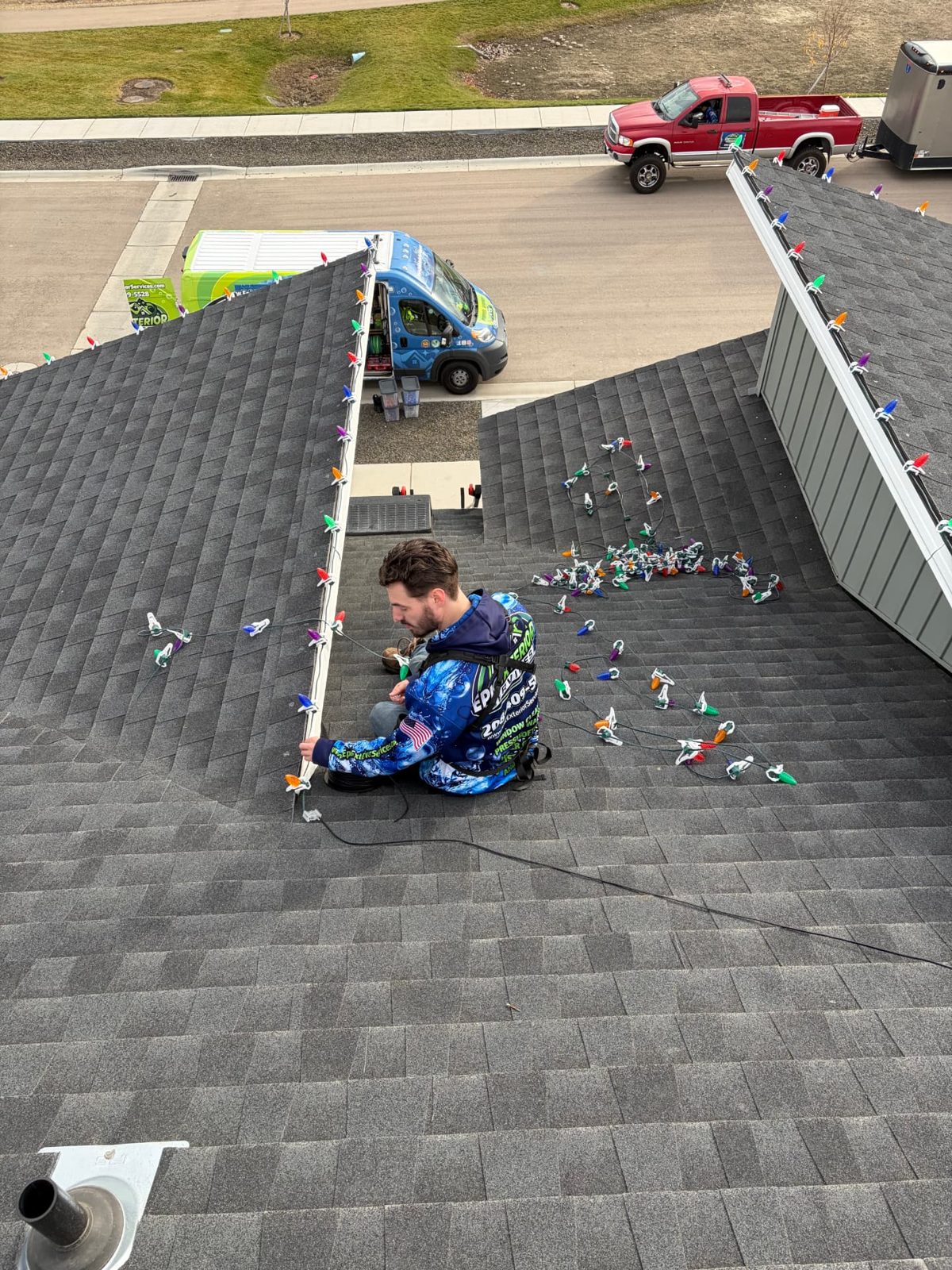 Installer clipping multicolor C9 lights along a roof valley with the Epic Exterior Services van parked below